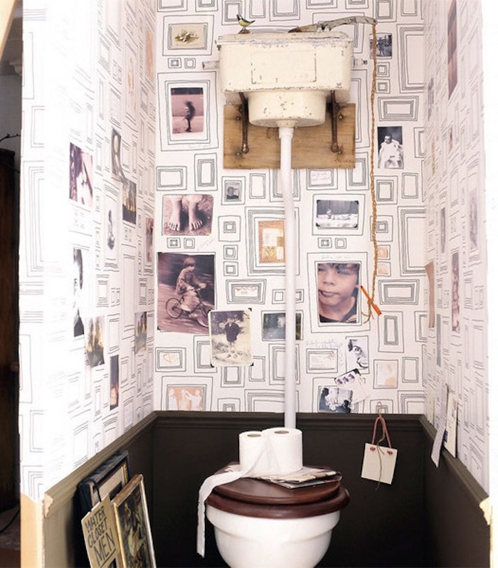 small toilet with dark grey panelling, white wallpaper with many drawn frames, some decorated with black and white photos, plain white toilet seat, with dark wooden cover