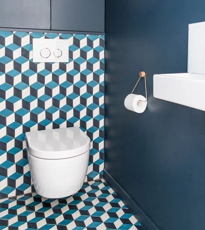 bathroom remodel, toilet with dark blue wall, other visible wall and floor have an unusual covering with geometric, faux 3D patterns in white, dark and light blue, modern white toilet seat and sink