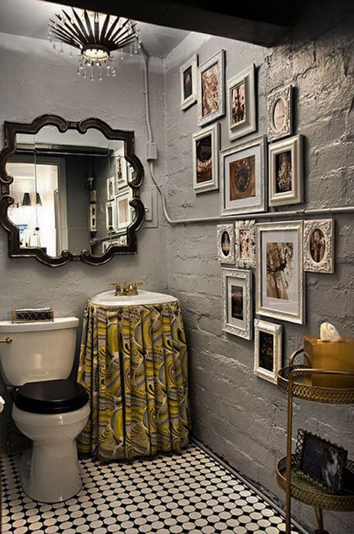 toilet with rough walls painted in grey, with a large baroque wall mirror, one wall decorated with many photos in different white frames, white and black tiled floor, classic toilet seat, and sink with yellow and grey patterned curtain
