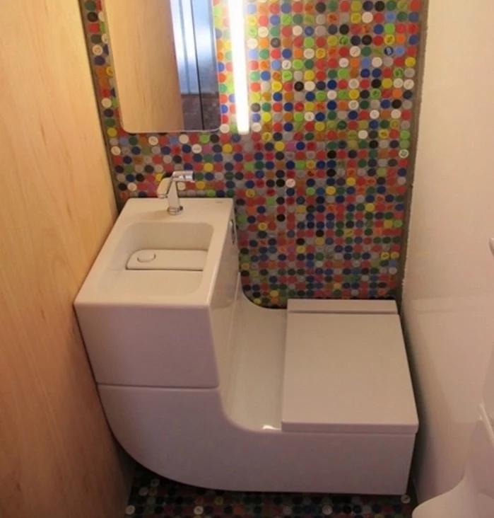 bathroom remodeling, small space with white sink, and toilet seat all in one, one wall is covered in pale wooden board, one is white, and one is decorated with multicolored mosaic, with mirror and light 