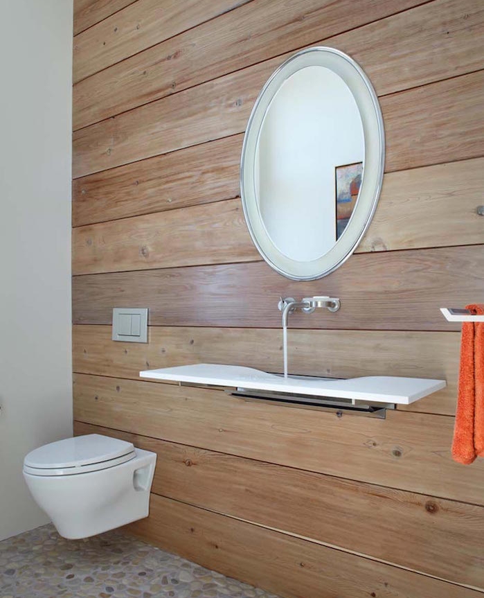 bathroom remodels, spacious toilet with one wall covered in wooden planks, other visible wall is white, round mirror with silver frame, white minimalist modern sink, pebbled floor and plain white toilet seat