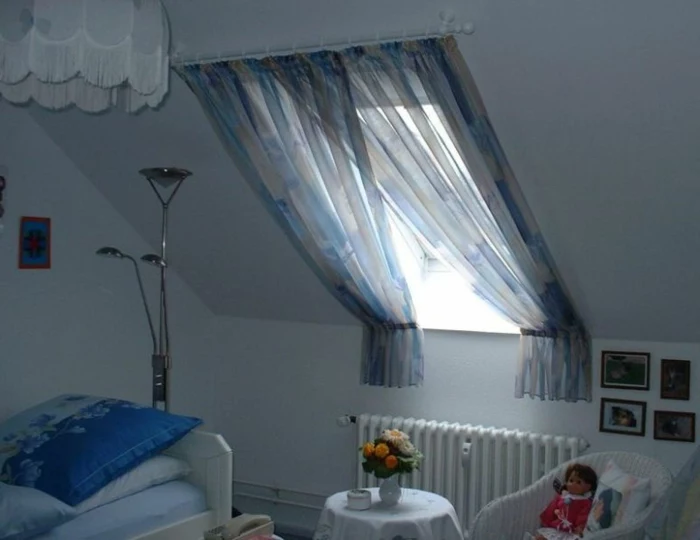 room with bed in pale and dark blue, small white round table and chair, sloping window with overlapping curtains, in pale blue and grey