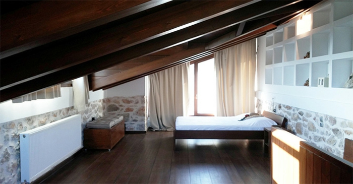 bedroom curtains, sloping ceiling with wooden beams, stone paneling and white shelves, wooden bed and floors, large window with cream-colored curtains
