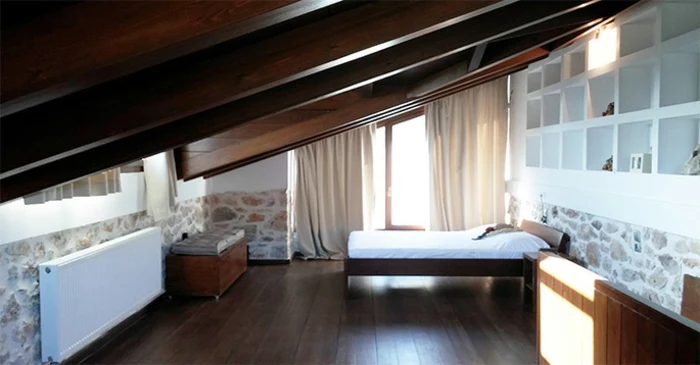 bedroom curtains, sloping ceiling with wooden beams, stone paneling and white shelves, wooden bed and floors, large window with cream-colored curtains