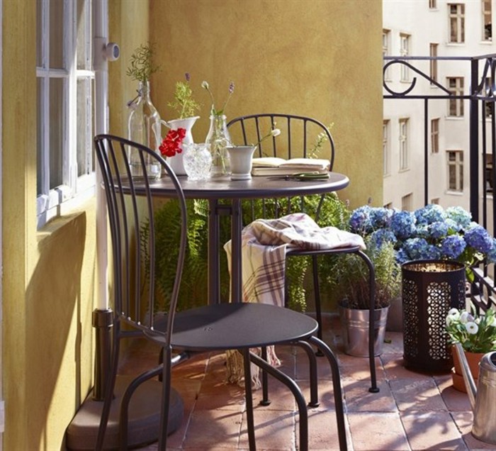 black round table with two matching chairs, near yellow wall, potted plants and black railing, porch décor