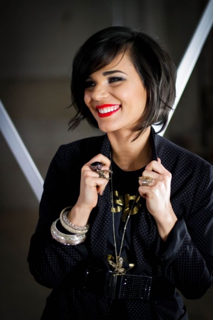 woman with red lipstick and big smile, wearing a black shirt with polka dots, and several pieces of jewelry, black side parted pageboy haircut