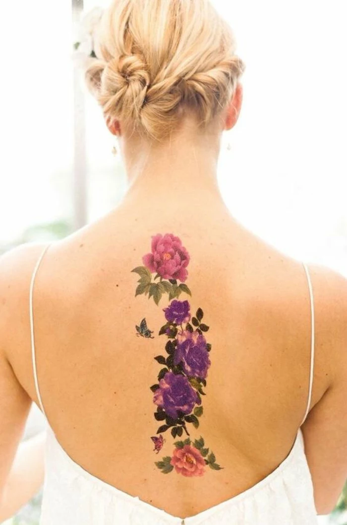 traditional flower tattoo, blond woman with hair put up, wearing a white open back top, revealing a large, vertical tattoo of a pink peony, above several violet roses, and a single pink rose, many dark and light green leaves