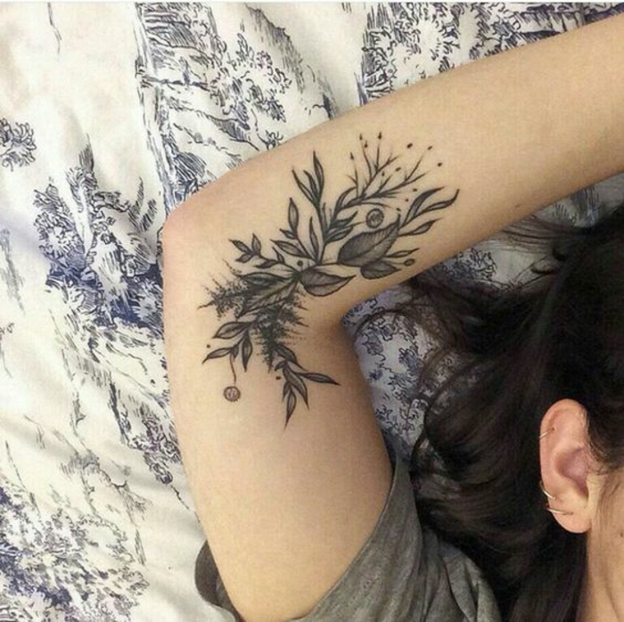close up of a woman's arm, with a dark tattoo near her elbow, depicting various different leaves and blossoms