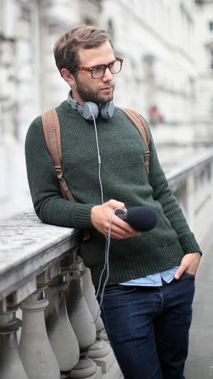 green woolen jumper, over pale blue shirt, and dark blue jeans, casual clothes for men, worn by young man with glasses, with brown backpack, and grey headphones