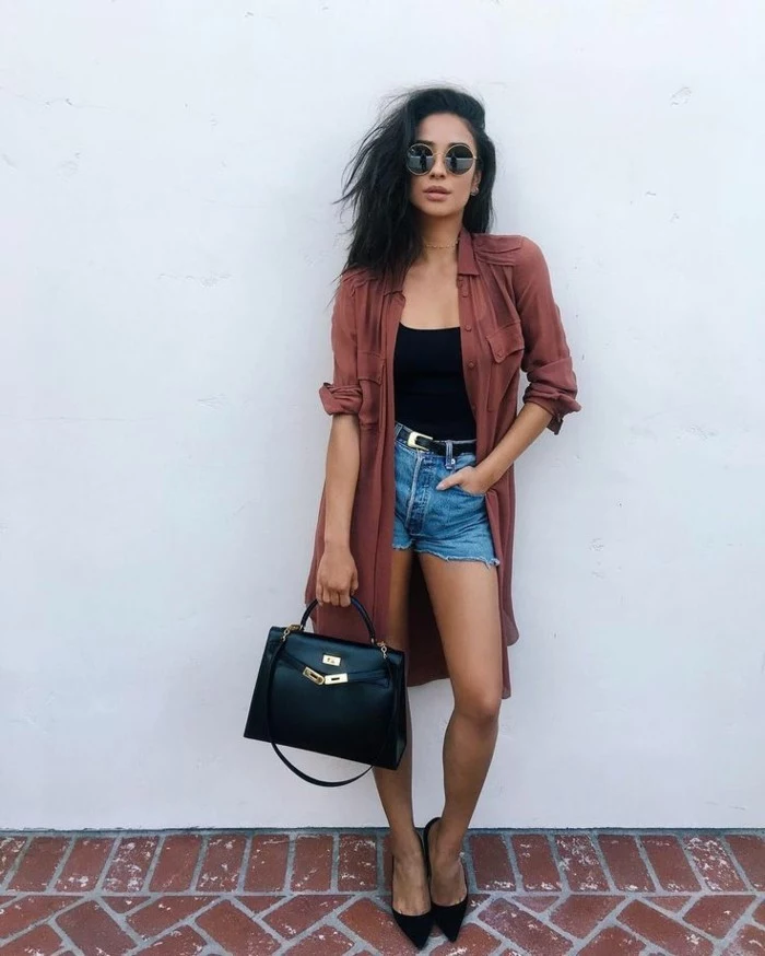 women outfits, black-haired woman with round sunglasses, wearing pale red oversized shirt with rolled sleeves, over black top and cut off denim shorts, with black belt, high heels and black leather bag