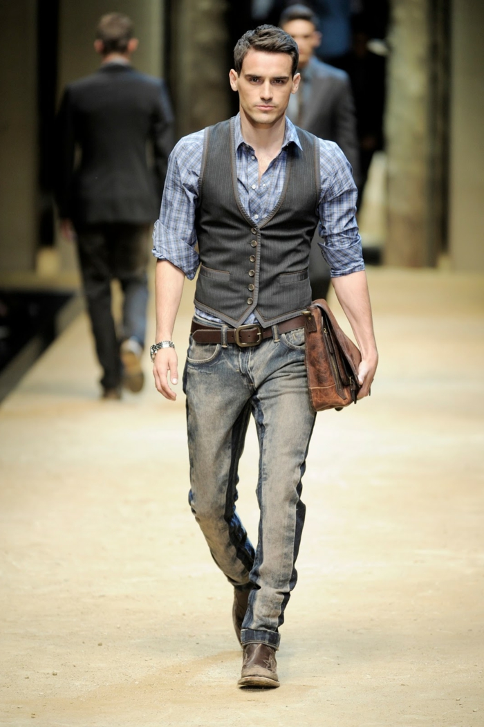 male catwalk model, wearing pale blue chequered shirt, grey button up vest, distressed cowboy jeans, and worn vintage shoes, casual clothes for men, antique looking suede bag in his hand