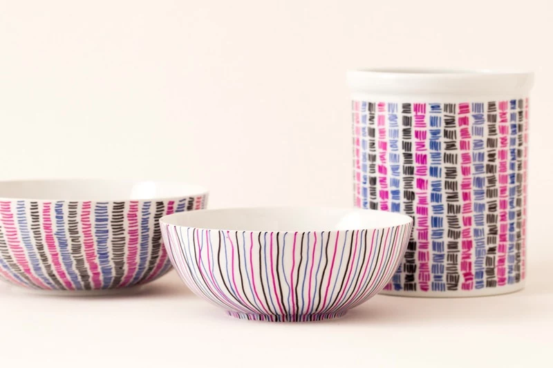 two white ceramic bowls, and a mug, decorated with lines, and tiny scribbles, in pink blue and black