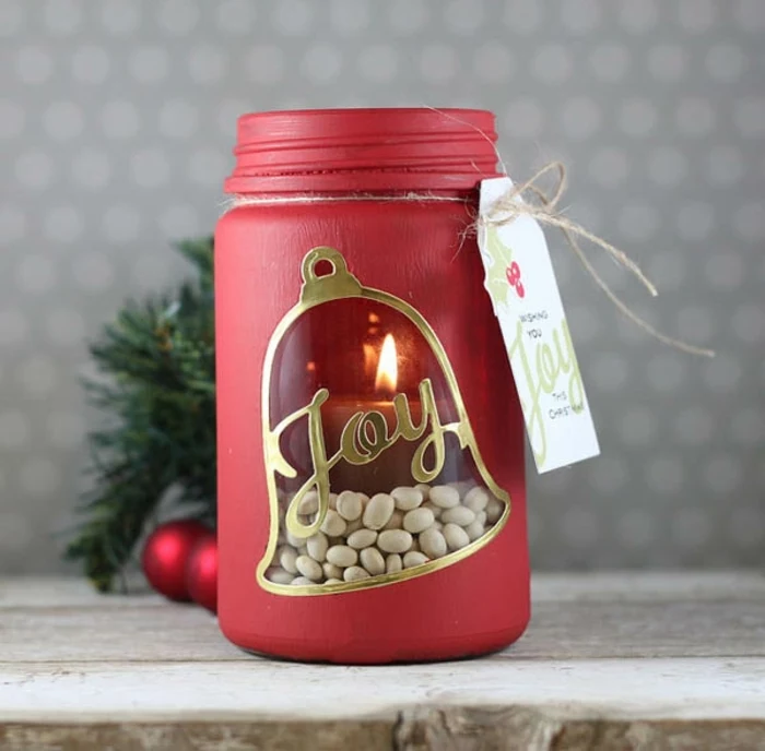 christmas mason jars, a jar painted in red, with a bell-shaped window, decorated with gold and the word joy, and revealing a lit candle, placed on a row of white beans