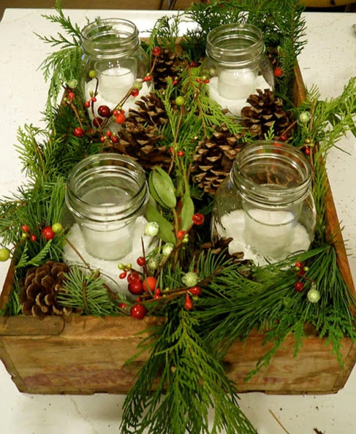 christmas mason jars, four jars containing fake snow, and small glasses with candles, placed in a wooden tray, next to fir branches, fir cones and berries, and other plants