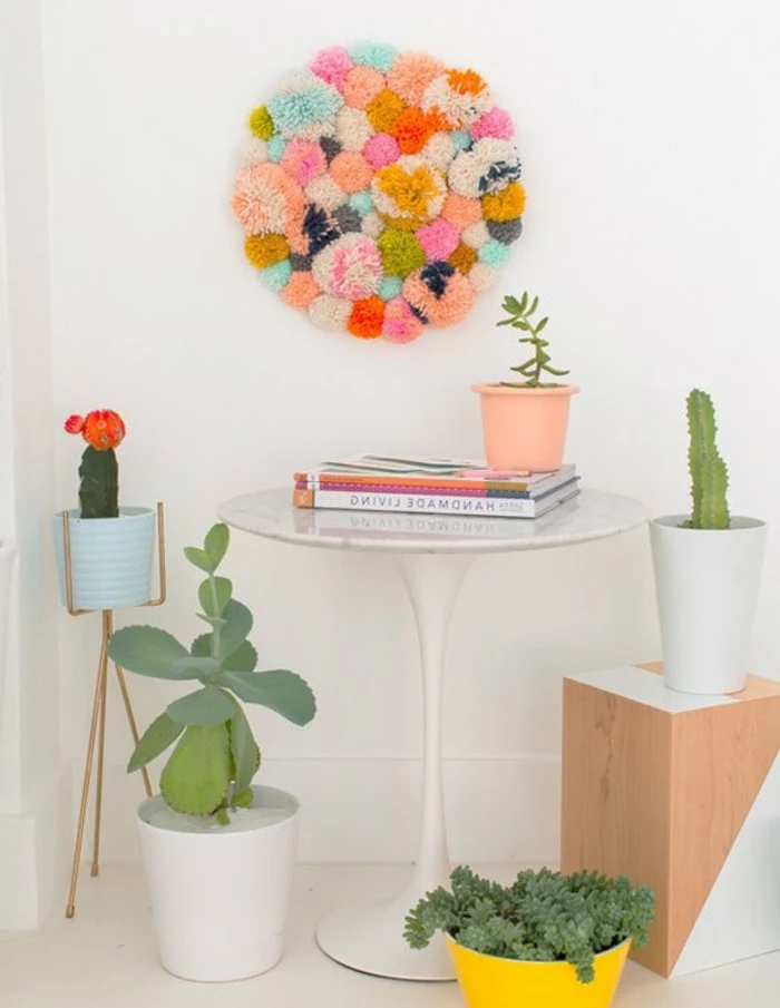 round wall decoration, made from multicolored pom-poms stuck together, easy arts and crafts, hanging over a small white table, with wooden stool, and potted plants