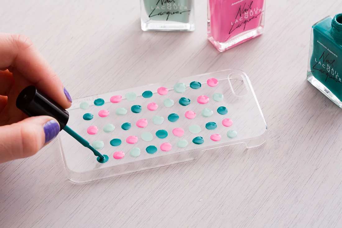 homemade crafts, woman's hand with purple nails, decorating a clrear phone case, with dots of pale blue and pink, and teal nail polish