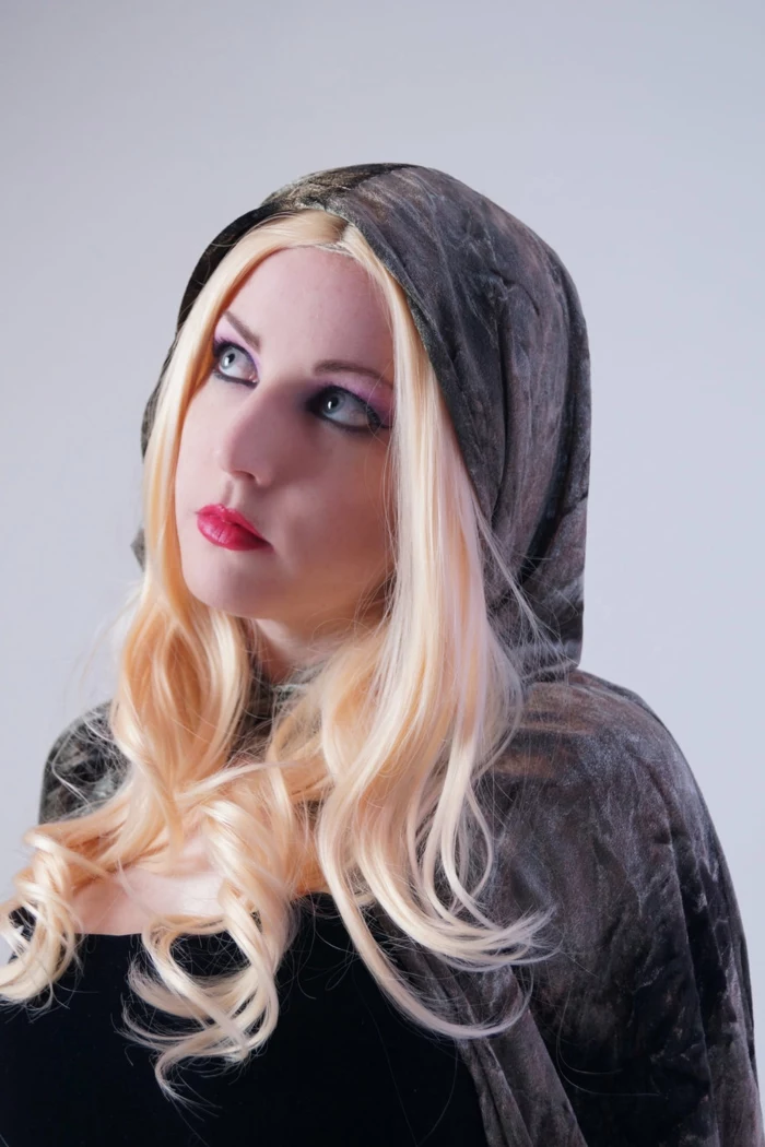 woman with red lipstick, black eye-make-up and blonde wig, wearing a black top, and dark grey velvet cape and hood, renaissance braided hairstyles