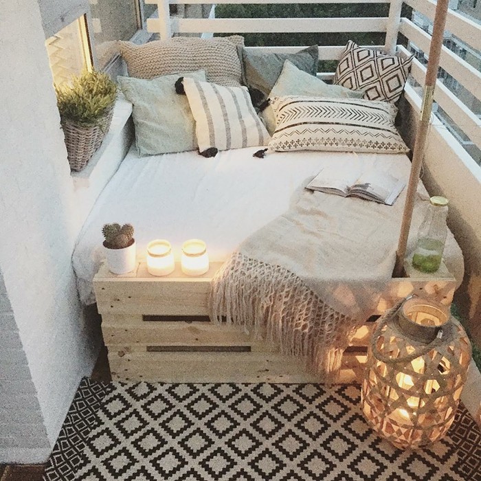 covered-patio-ideas-settee-made-from-wooden-pellets-with-white-cover-and-many-patterned-pillows-in-pale-pastel-colors-near-dark-grey-and-cream-patterned-rug-pale-beige-throw-and-large-wicker-lantern porch ideas, settee made from wooden pallets, with white cover, and many pale patterned cushions, several lit candles, potted plants and blanket