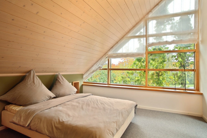 bedroom with sloped ceiling, covered with wooden planks, asymmetrical window with short, sheer asymmetrical curtain, pale brown bed
