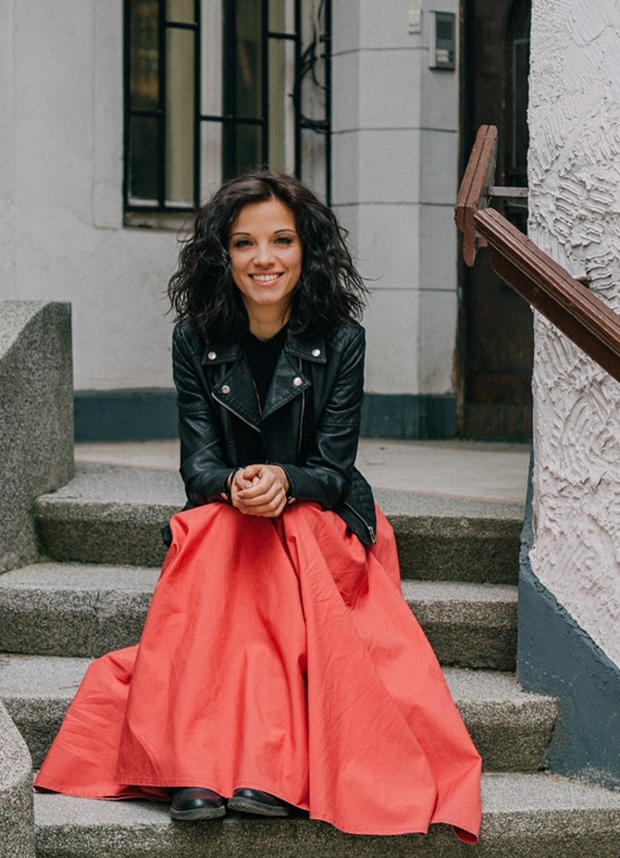 casual clothes, smiling woman with curled brunette hair, sitting on stone steps, and wearing coral red maxi skirt, black top and black leather jacket