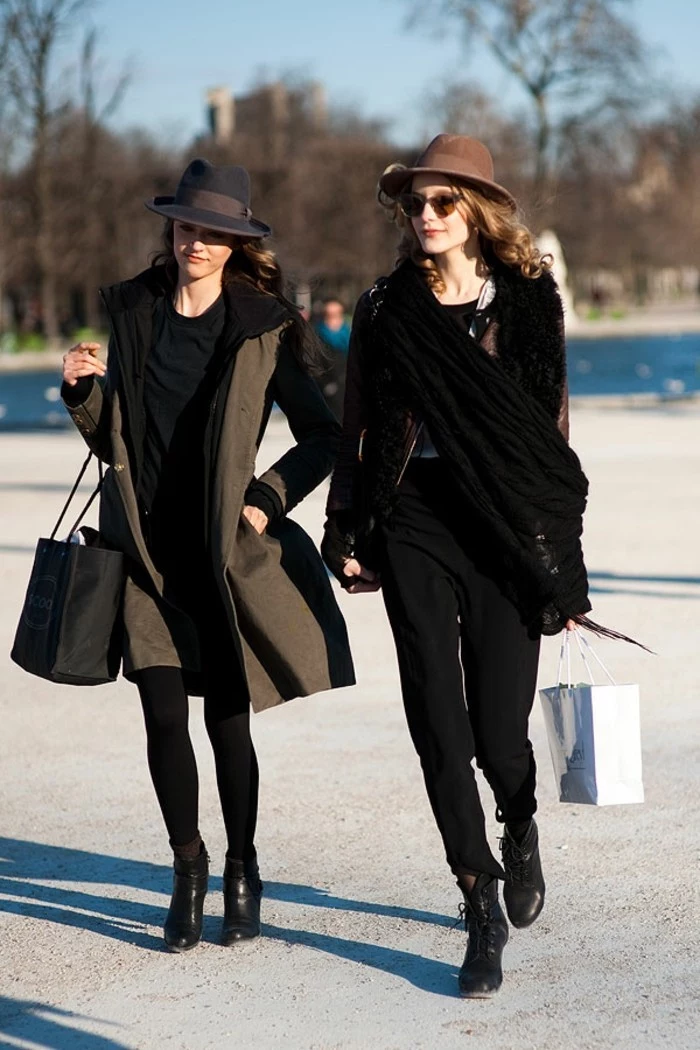 women business casual, woman in black outfit, with grey winter coat and black felt hat, walking next to woman in black trousers, with brown felt hat, and black scarf