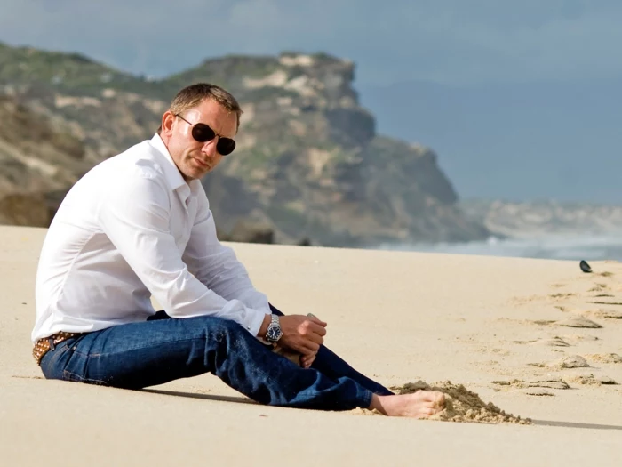 skinny business casual jeans, and white shirt, worn by daniel craig, with aviator sunglasses, sitting barefoot, on a sandy beach 