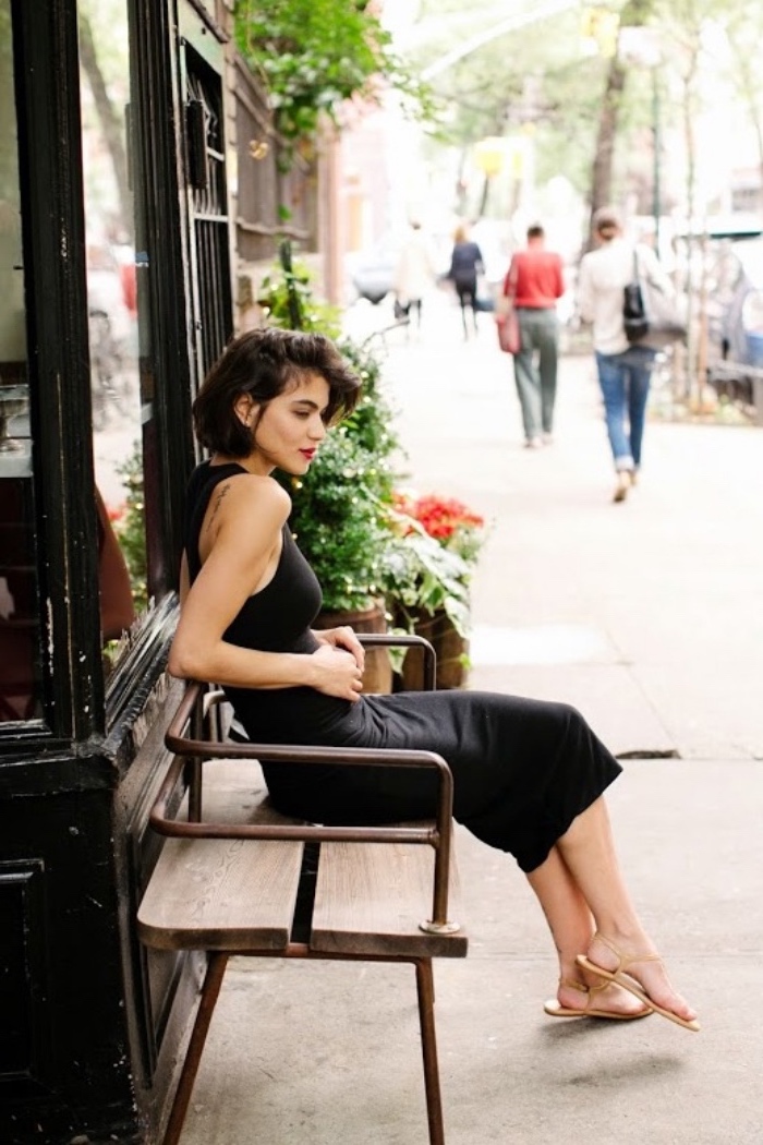 brunette woman with short bob haircut, wearing black midi dress, and beige sandals, and sitting on a bench
