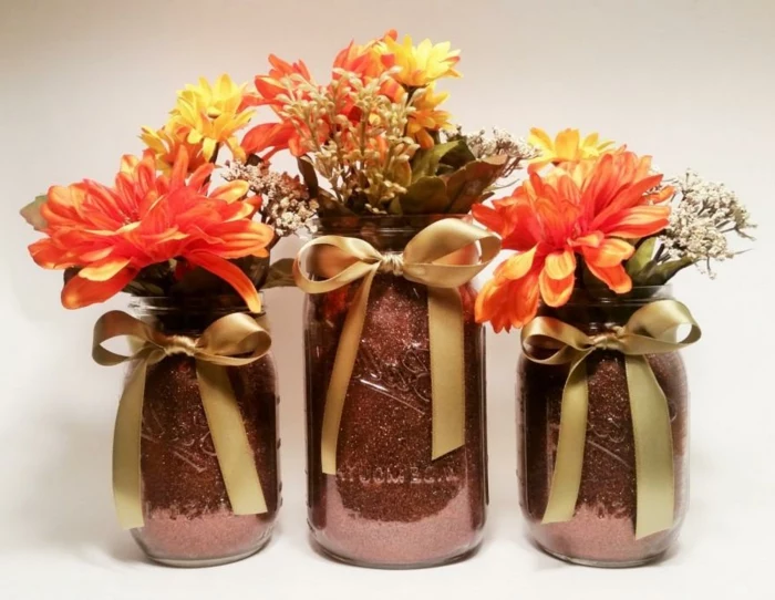 diy mason jar, three jars of different sizes, painted in a dark pink, glittery color, and decorated with gold bows, containing orange and yellow flowers