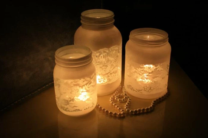 diy mason jar, three differently sized jars, painted white and featuring a lace pattern, lit from within and placed in a dark room, near a pearl necklace