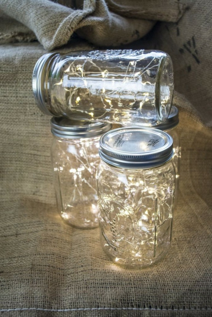 mason jar centerpieces, four mason jars, placed on burlap cloth, with closed aluminium lids, each containing lit fairy lights