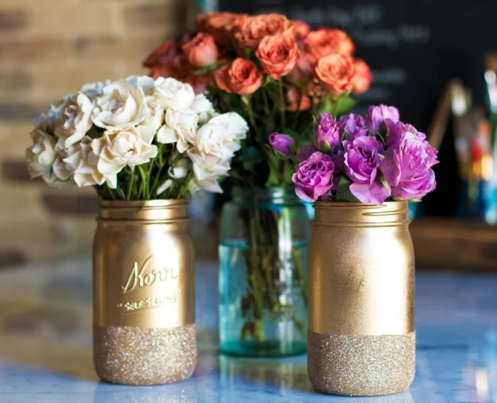 mason jar crafts, two jars spray-painted in gold, with glitter details, containing white and purple flowers, and a clear blue jar, with orange roses, in the background
