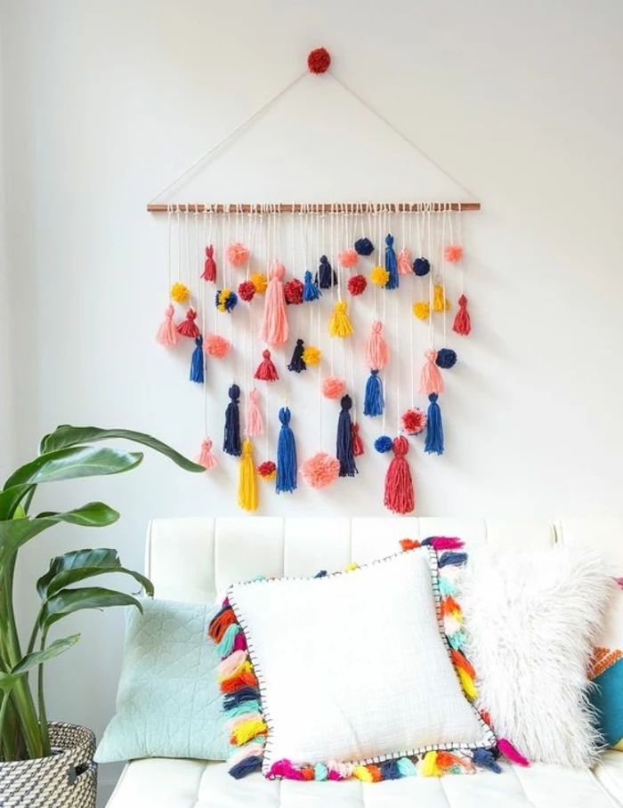 wall decoration made from a piece of wood, adorned with pom-poms, in different shapes and colors, homemade crafts, hanging over a white couch