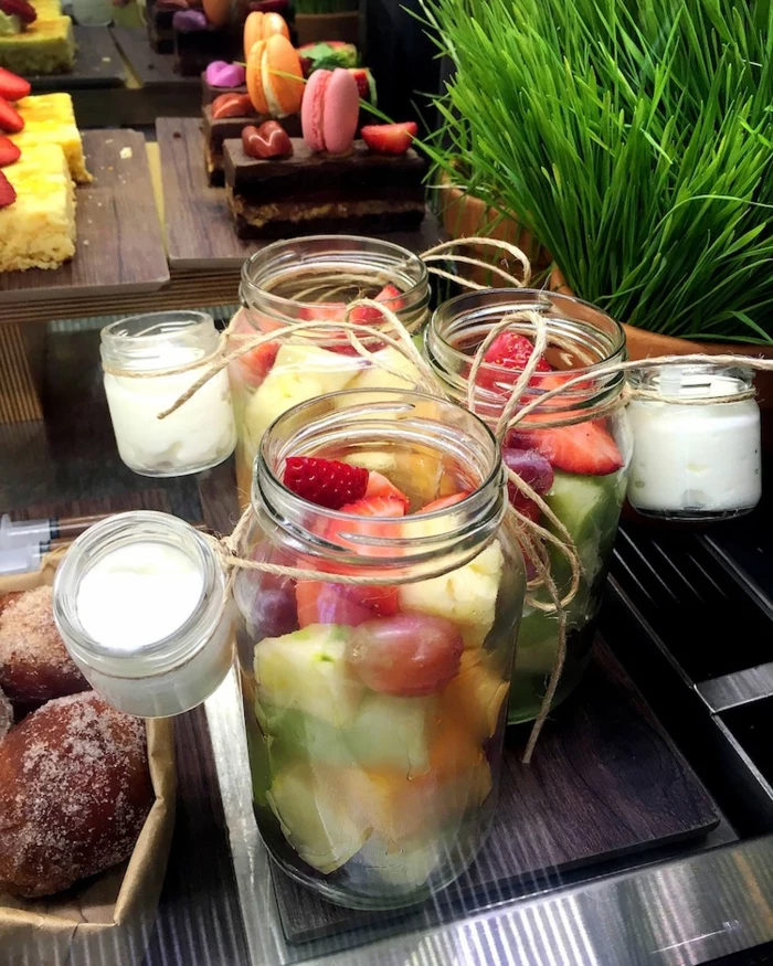 mason jar gifts, three jars containing fruit salad, with smaller jars, filled with cream, tied to them with string, cakes and pastries in background