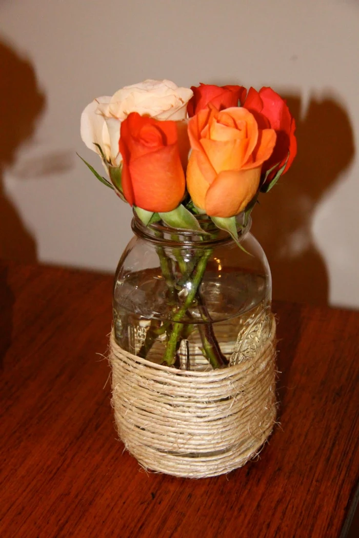 mason jar wrapped around with string, containing four orange roses, and one white, placed on a wooden table