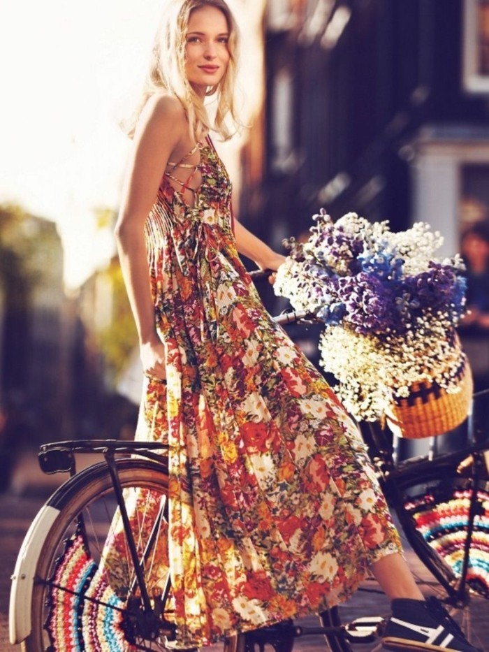 smiling blonde woman, wearing multicolored floral maxi dress, and black and white sneakers, on bike with basket, containing white blue and purple flowers