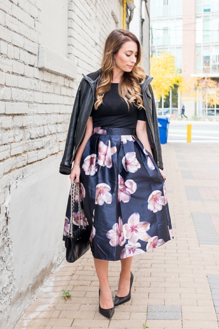 dress attire, shiny dark blue midi skirt, with pale pink flowers, worn with black semi-sheer t-shirt, by woman with honey blonde wavy hair, with black leather jacket over her shoulders