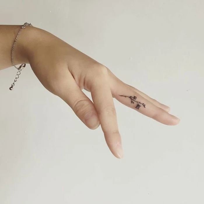 wildflower tattoo, close up of a female hand, with delicate silver bracelet, a tattoo of a tiny minimalistic flower, in black ink, on t6he side of her middle finger