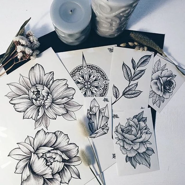 flower tattoo designs, several sheets of white paper, with various tattoo designs, four flowers and a leafy plant, a mandala and a crystal, small dried flowers and candles nearby