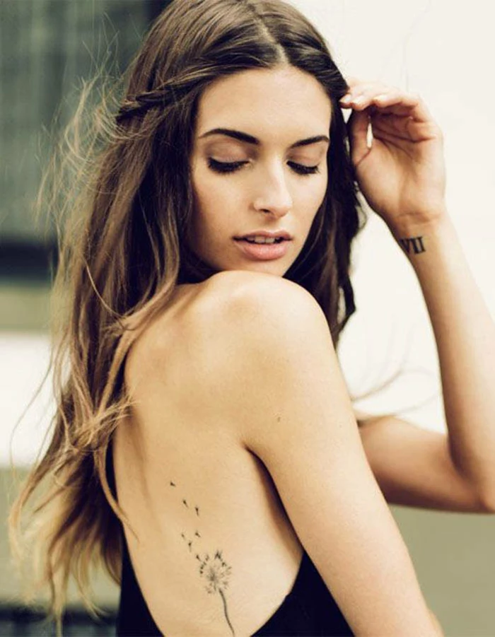 flower tattoos, brunette with long wavy hair, wearing a black top with open back, looking over her shoulder, a black tattoo of a dispersing dandelion, on her lower back, VII tattooed on her wrist