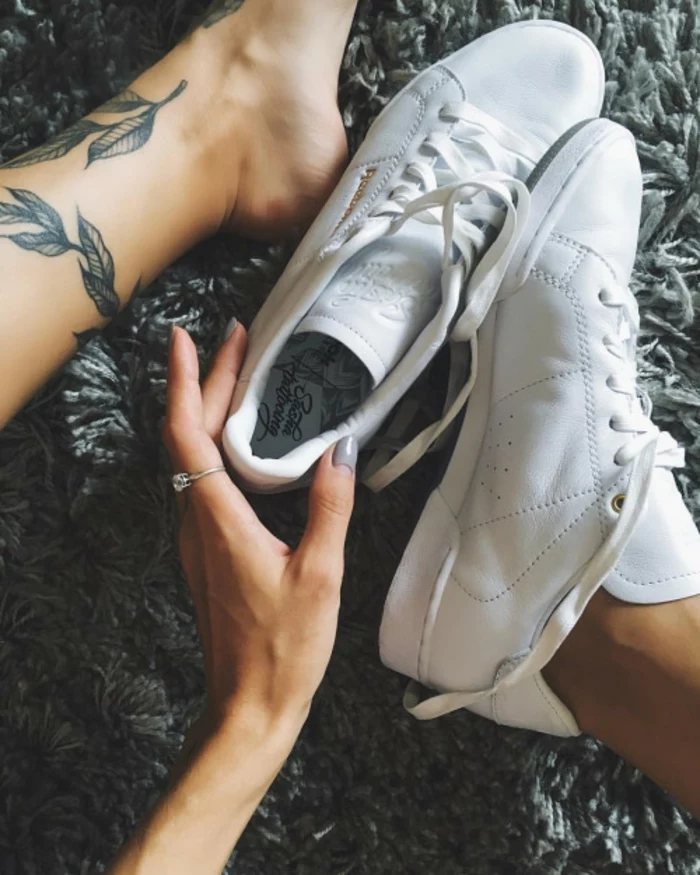 flower tattoos, close up on woman's feet and hand, one bare foot, with black ink leaf tattoos, other one in a white leather sneaker, hand holding other sneaker