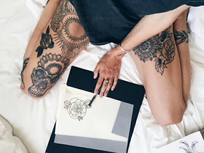 flower tattoos, woman in black top, with various tattoos on her legs, sitting on a bed, holding a pen, near a piece paper, with an outlined drawing of a rose