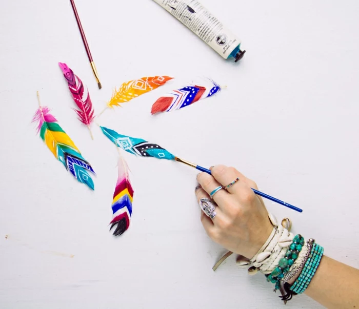 feathers painted in different colors, and decorated with patterns, homemade crafts, hand with many bracelets and rings, holding a blue brush
