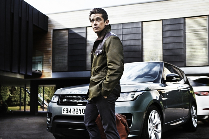 khaki winter jacket, with black leather details, combined with dark grey trousers, business casual outfits, worn by man holding brown travel bag
