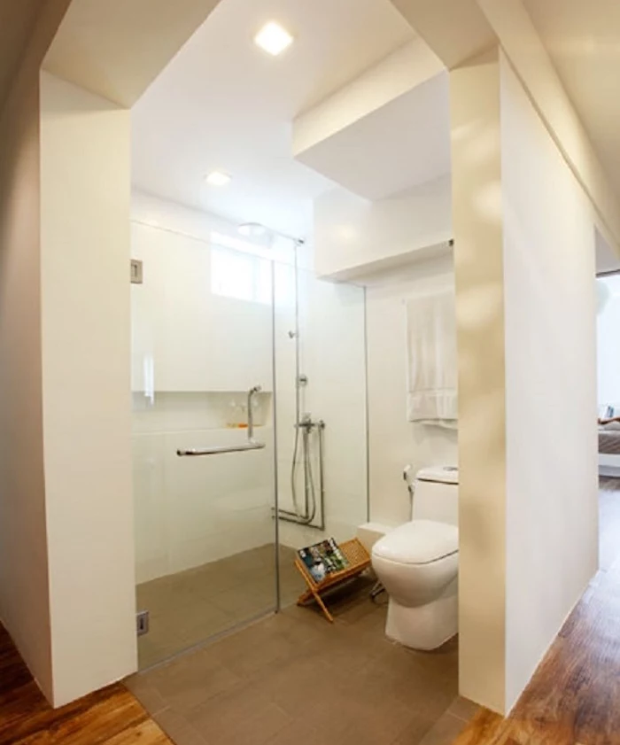 spacious semi-detached open plan bathroom, with thick white walls, containing large glass shower cabin, modern oval toilet seat, brown carpet and wooden floor