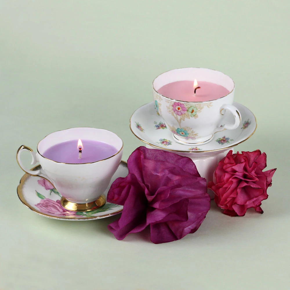 lit candles in pale pink and violet, inside porcelain tea cups, placed on saucers, fun and easy crafts, two purple flower ornaments nearby