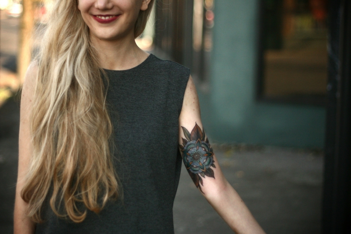 traditional flower tattoo, smiling woman with long blonde hair, red lipstick and dark grey top, with a blue flower mandala tattoo, resembling the tudor rose
