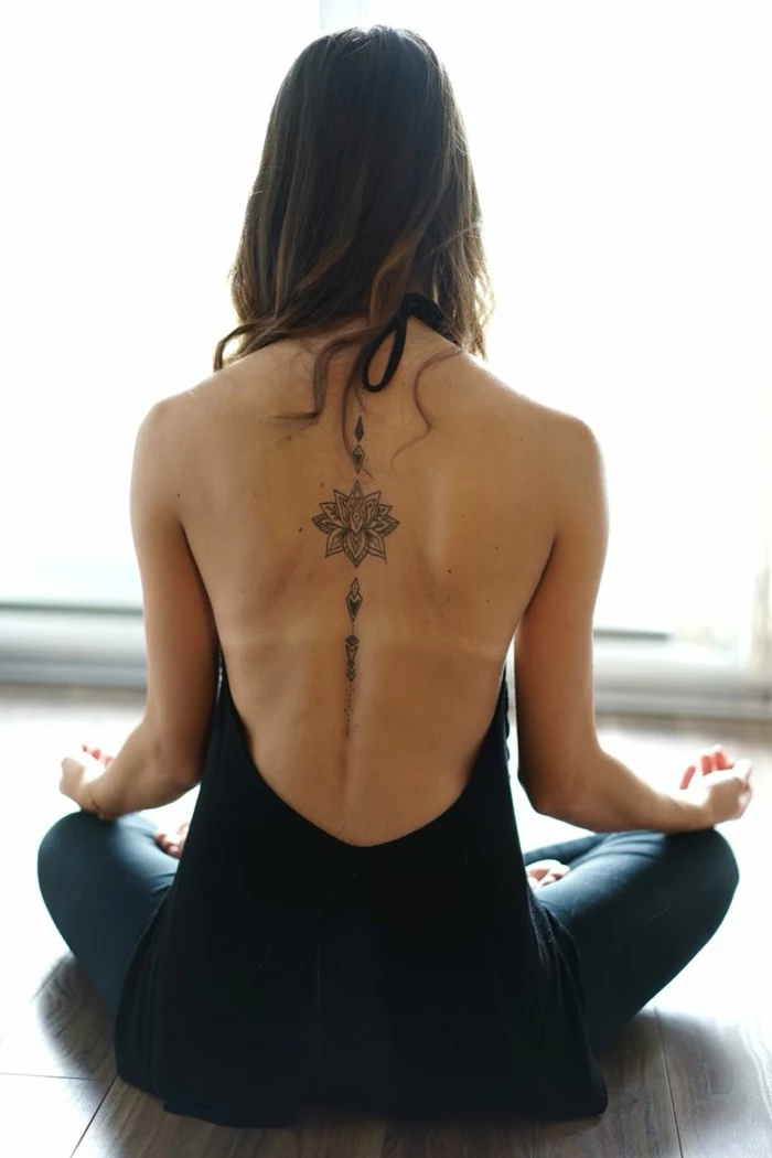 lotus flower tattoo, brunette woman sitting in lotus position, with an open back black top, and blue jeans, with lotus tattoo, outlined with black in on her back