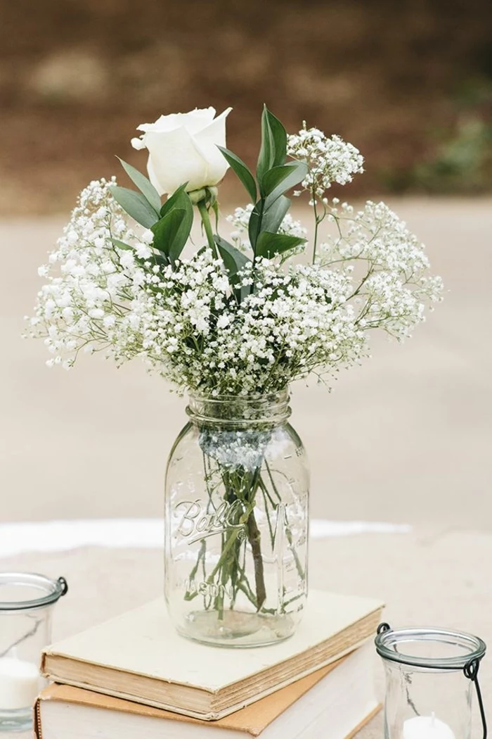 mason jar centerpieces, a clear mason jar, placed on stacked beige books, containing a single white rose, green leafy plant, and many tiny white flowers