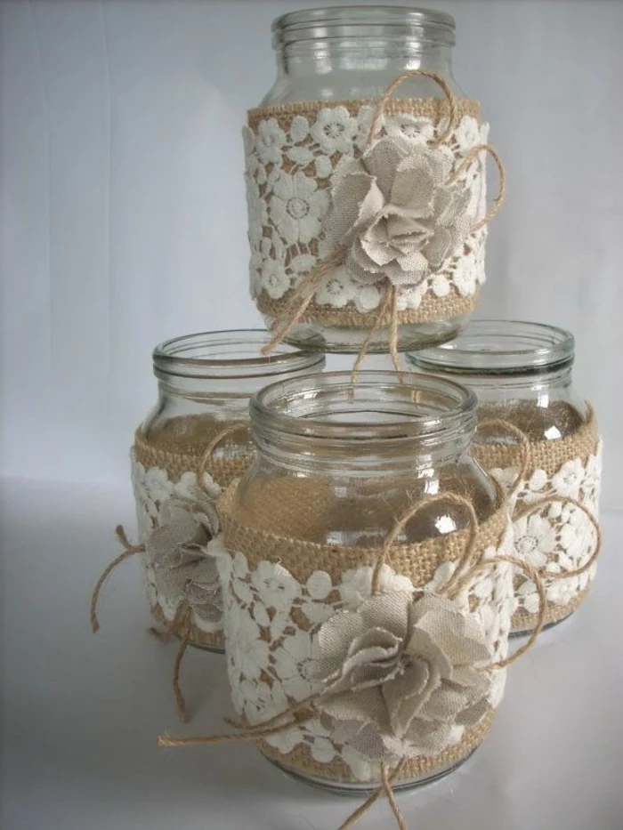 mason jar centerpieces, four identical jars, decorated with white floral lace, placed on top of beige burlap, with burlap flowers and string bows