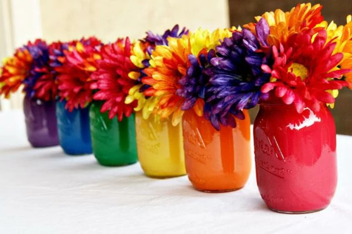 six mason jars, each painted in a different color, purple and blue, green and yellow, orange and pink, containing mixed flowers, in the same colors